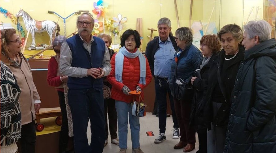 Varios compoñentes do grupo do Fogar Parroquial Santa María e Santiago da Coruña forman un corro ante a explicación de Ricardo Pérez y Verdes diante dun expositor no que se ve un cabaliño de xoguete.